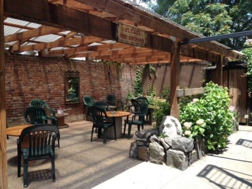 Outdoor seating area with wooden beams, green chairs, and tables surrounded by plants and a stone feature.