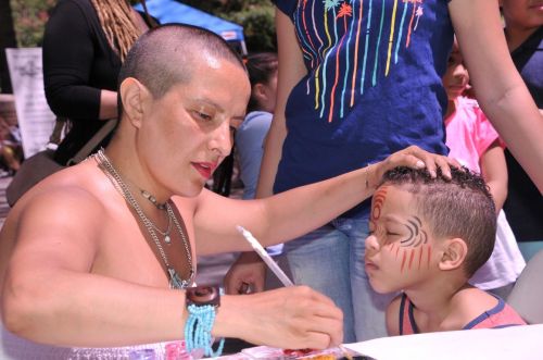 A woman paints a design on a child's face at an outdoor event, surrounded by people and colorful decorations.