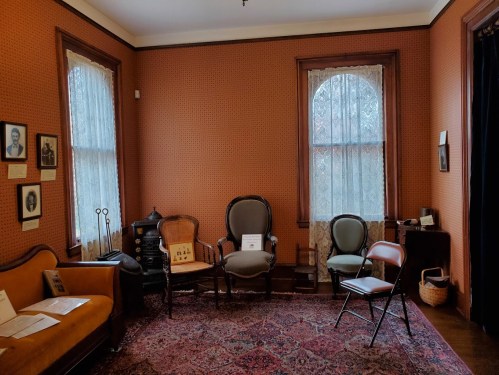 A cozy room with vintage furniture, including chairs and a sofa, adorned with patterned wallpaper and large windows.