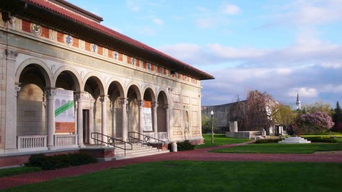 A historic building with arches and banners, surrounded by a green lawn and trees under a partly cloudy sky.