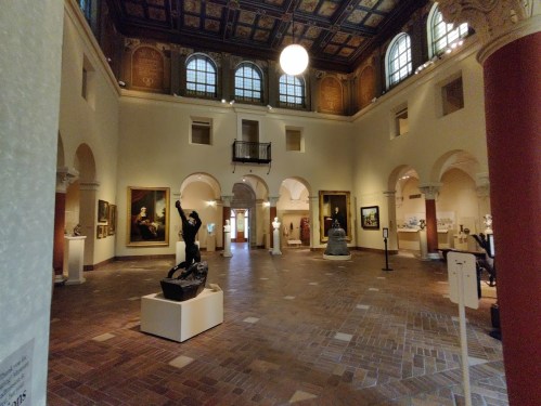 Interior of an art gallery featuring sculptures and paintings, with arched windows and a high ceiling.