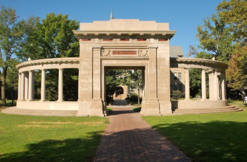 A stone archway with columns, surrounded by greenery and a pathway leading to a building in the background.