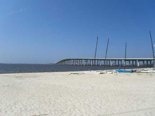 A sandy beach with sailboats and a long bridge extending over calm water under a clear blue sky.