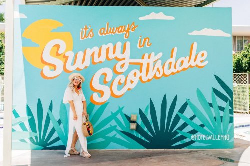 A woman in a white outfit poses by a colorful mural that says "It's always Sunny in Scottsdale."
