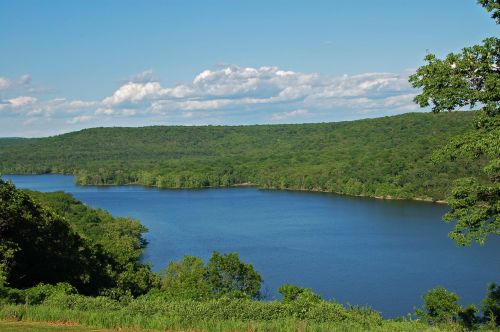 A serene lake surrounded by lush green hills under a clear blue sky with scattered clouds.