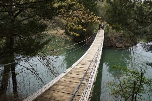 A wooden suspension bridge spans a calm, green river, surrounded by trees and natural scenery.