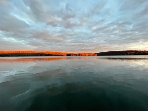 A serene lake at sunset, reflecting colorful clouds and a tree-lined shore under a dramatic sky.