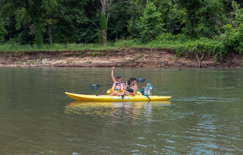 Two people in a yellow kayak on a calm river, surrounded by lush greenery, one waving at the camera.