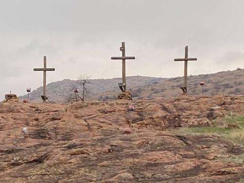 Four wooden crosses stand on rocky terrain, with a cloudy sky in the background.