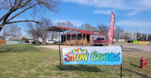 A colorful snow cone stand with a banner, truck parked nearby, and trees under a blue sky.