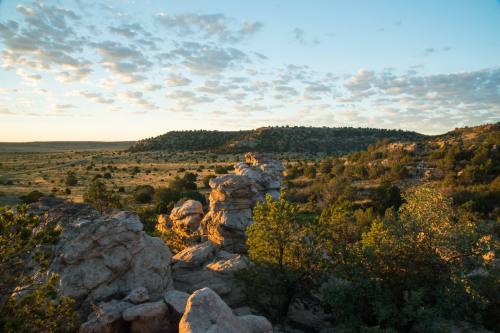 A scenic landscape featuring rocky formations, green shrubs, and rolling hills under a blue sky with scattered clouds.