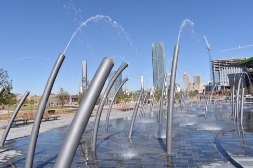 A modern fountain with tall, curved spouts spraying water, set against a city skyline on a clear day.