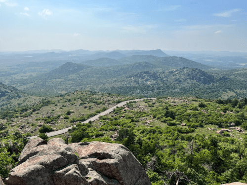 A scenic view from a rocky overlook, showcasing rolling hills and a winding road through lush green landscape.