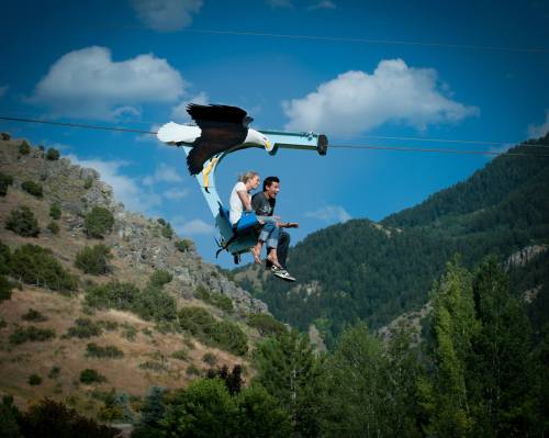 Two people ride a zip line with a large eagle sculpture soaring above them, set against a mountainous landscape.