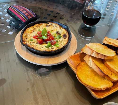 A skillet of baked cheese topped with tomatoes and herbs, alongside toasted bread and a glass of red wine.