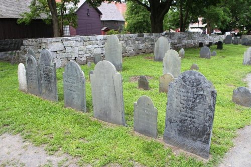 A historic cemetery with weathered gravestones set in a grassy area, surrounded by stone walls and trees.