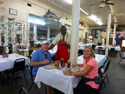 A smiling waiter in an apron poses with two diners at a restaurant table, enjoying their meals in a casual setting.