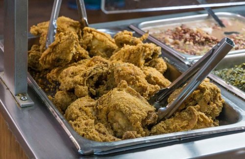 A metal tray filled with golden-brown fried chicken pieces, with tongs resting on the side.