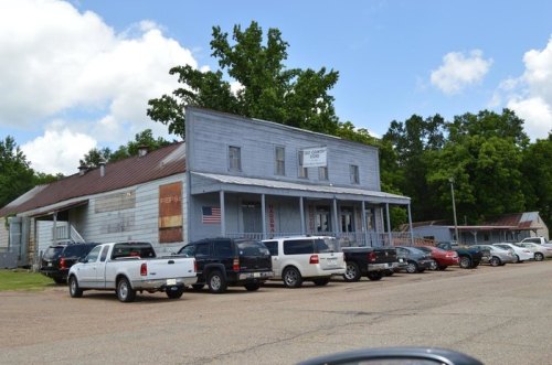 A weathered building with a sign, surrounded by parked cars, set against a backdrop of trees and a blue sky.