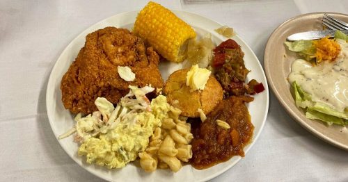 A plate of fried chicken, corn on the cob, coleslaw, macaroni, cornbread, and baked beans on a white tablecloth.