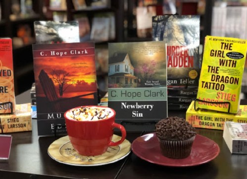 A cozy display of books by C. Hope Clark, a red mug of hot chocolate, and a chocolate cupcake on a plate.