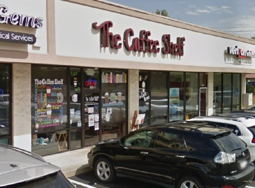 Exterior view of "The Coffee Shelf" shop with large windows displaying colorful signs and a parked black SUV in front.