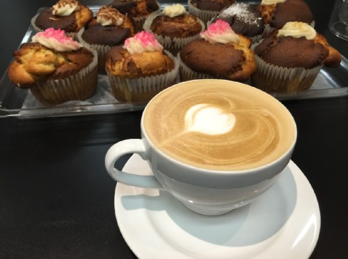 A latte with a heart design in a white cup, with a tray of colorful cupcakes in the background.