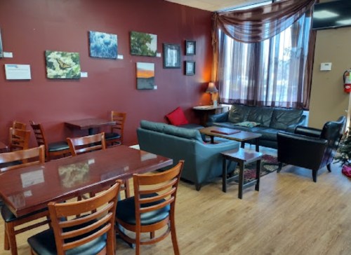 Cozy café interior with wooden tables, chairs, and a seating area featuring artwork on the walls and large windows.
