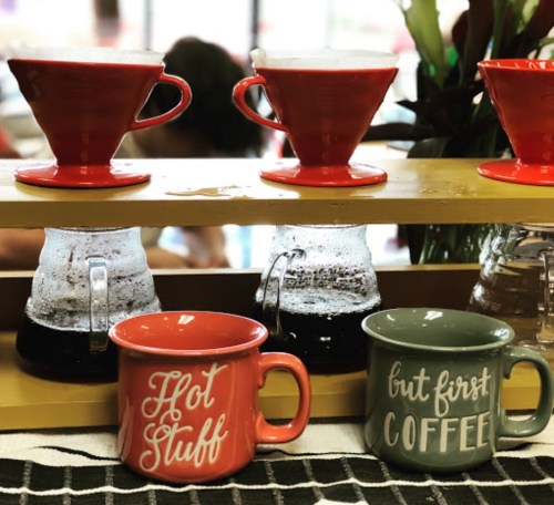 Two colorful coffee mugs sit on a shelf with brewing coffee in the background, one mug reads "Hot Stuff" and the other "But First, Coffee."