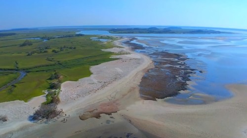 Aerial view of a coastal landscape featuring sandy shores, green marshlands, and calm blue waters.