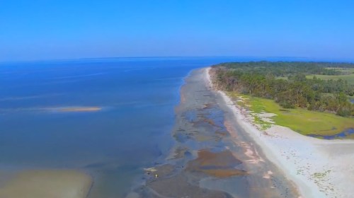 Aerial view of a serene beach with calm waters, sandy shore, and lush greenery in the background under a clear blue sky.