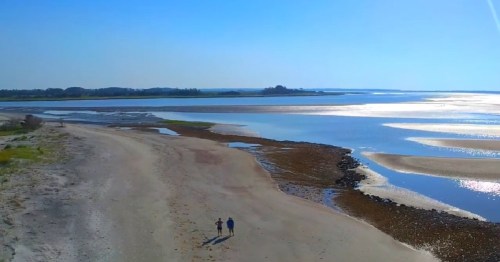 Two people walk along a sandy beach by a calm, reflective waterway under a clear blue sky.