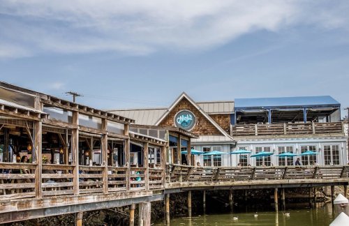 A waterfront restaurant with a wooden deck, outdoor seating, and a blue awning under a clear sky.