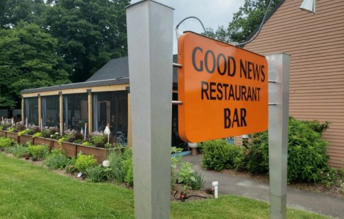Sign for "Good News Restaurant Bar" in front of a building with a garden and outdoor seating area.
