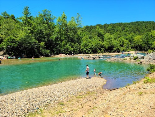 A sunny day at a clear, greenish-blue river with people enjoying the water and surrounding trees.