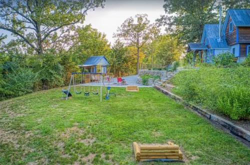A grassy backyard with a swing set, playhouse, and seating area surrounded by trees and a stone pathway.