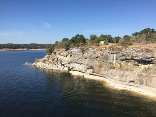 A rocky shoreline with a tent nestled among trees, overlooking a calm lake under a clear blue sky.