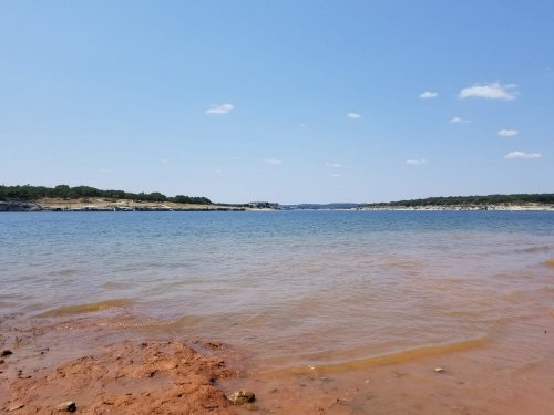 A serene lake view with clear blue water, sandy shore, and a bright blue sky dotted with a few clouds.