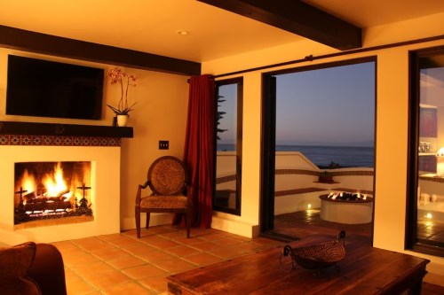 Cozy living room with a fireplace, large windows, and a view of the ocean at sunset. Warm and inviting atmosphere.