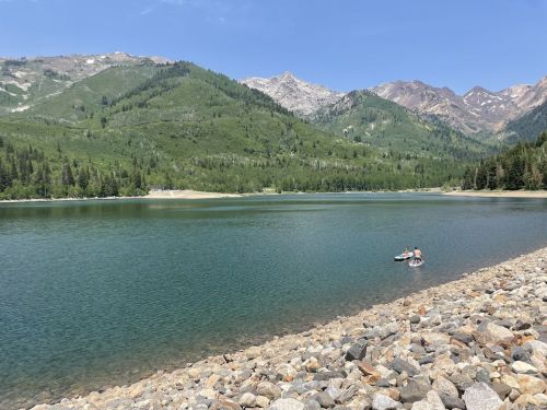 A serene lake surrounded by green mountains, with a rocky shore and two people on paddleboards in the water.