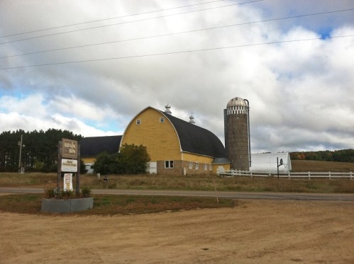 A yellow barn with a silo, surrounded by fields and trees, under a cloudy sky. A sign is visible in the foreground.