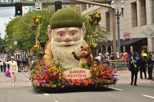 A large float featuring a giant gnome and a cartoon animal, decorated with colorful flowers, in a parade for a garden festival.