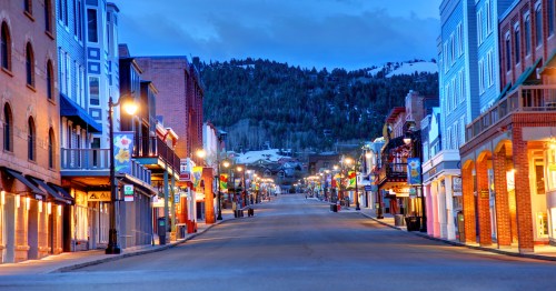 A quiet, empty street lined with colorful buildings, illuminated by streetlights, with mountains in the background at dusk.