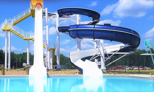A large water slide with blue and yellow sections towers over a clear swimming pool under a bright blue sky.