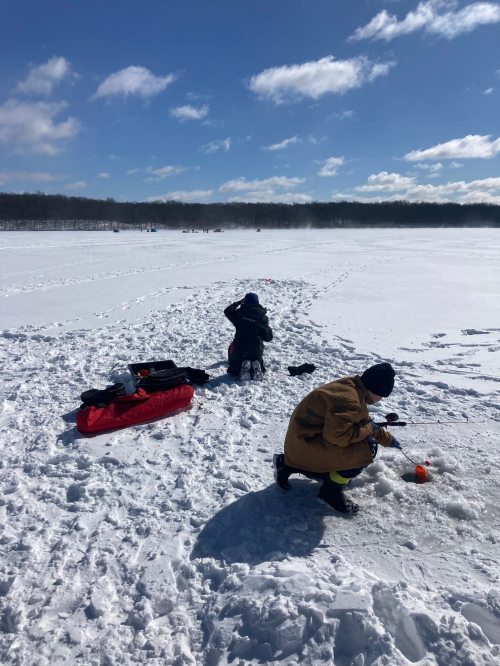 Two people ice fishing on a snowy lake, with a bright blue sky and distant trees in the background.