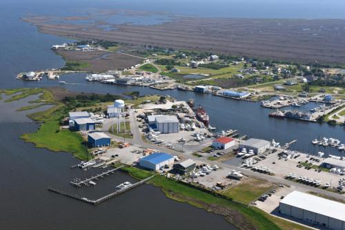 Aerial view of a coastal marina with boats, warehouses, and a water tower, surrounded by marshland and water.