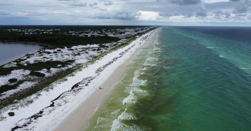 Aerial view of a sandy beach with turquoise waters, surrounded by greenery and a cloudy sky.