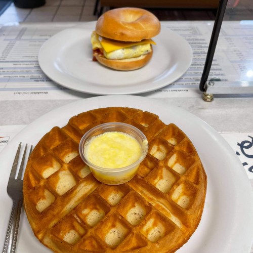 A plate with a waffle and a small cup of butter, alongside a sandwich on a separate plate.