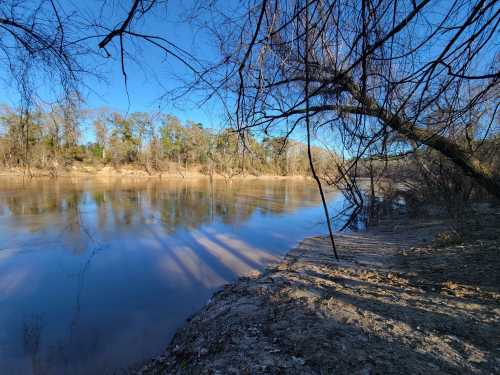 A serene river scene with calm water, sandy banks, and trees reflecting in the water under a clear blue sky.