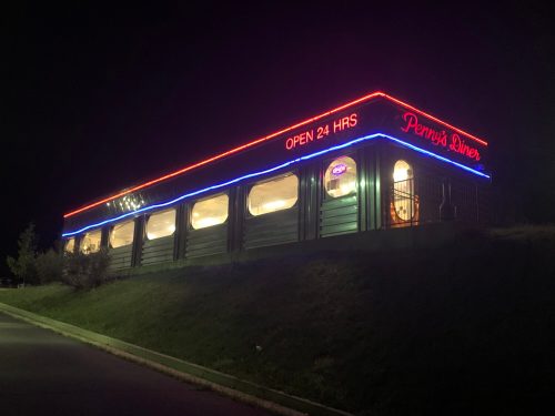 A brightly lit diner at night, featuring neon signs that read "Penny's Diner" and "Open 24 HRS."